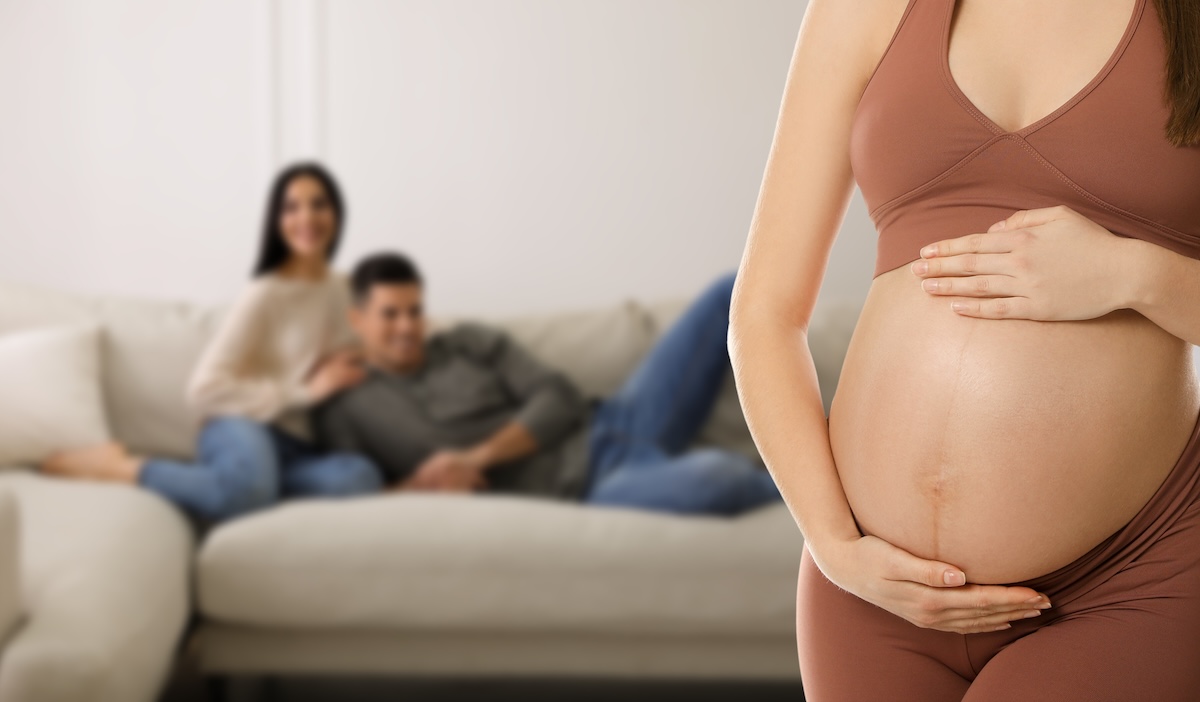 Pregnant woman in a sports bra holding her belly in the foreground, with a smiling couple sitting on a couch in the background, suggesting a surrogate relationship.