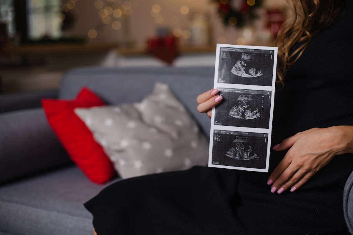 Close-up of a pregnant woman in a black dress holding ultrasound images on a couch, with festive lights softly glowing in the background.