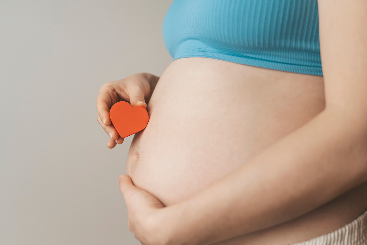 A pregnant individual in a blue top holding a red heart-shaped object against their bare belly.