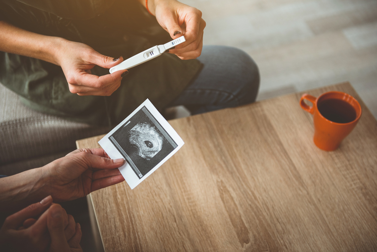 Dos manos, una sosteniendo una prueba de embarazo que muestra un resultado positivo y la otra sosteniendo una imagen de ultrasonido, con una taza sobre una mesa de madera al fondo.