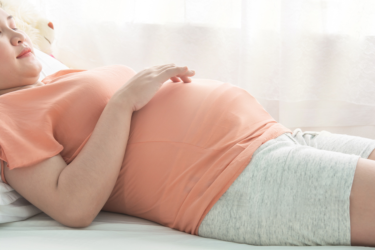 A pregnant individual lying down, wearing a peach-colored top and light grey shorts, resting their hand on their belly with a soft focus background.