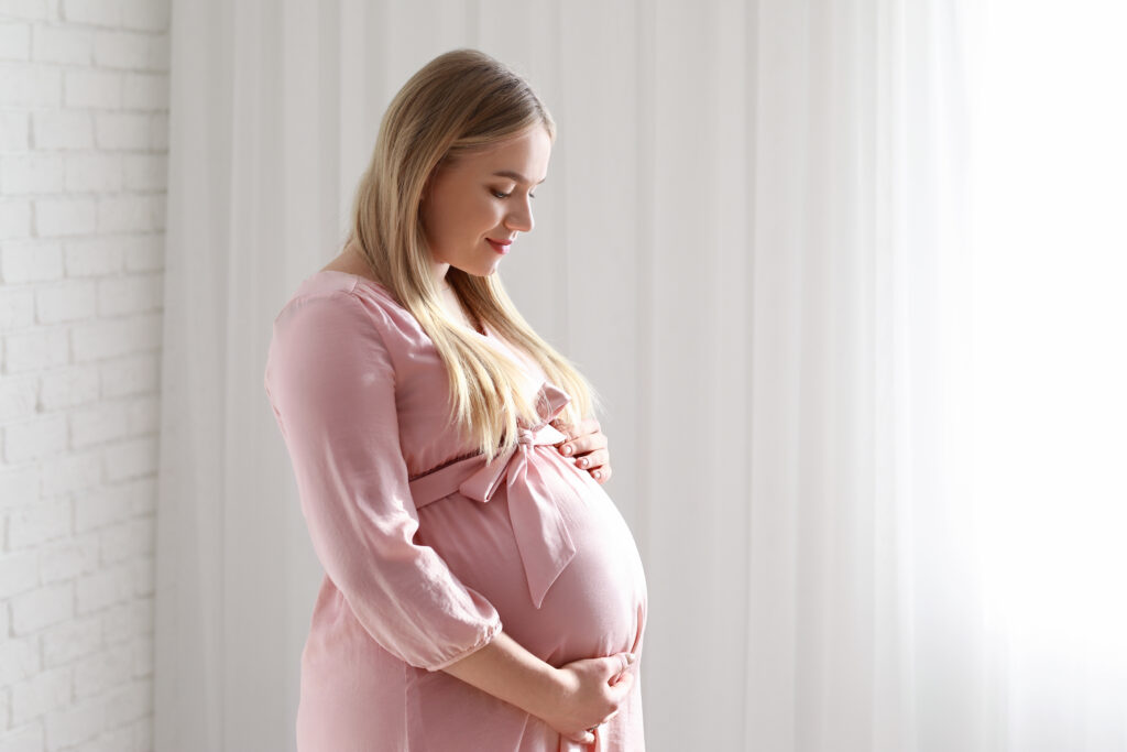 A pregnant woman in a pink dress gently cradling her belly, standing in a softly lit room with white curtains in the background.