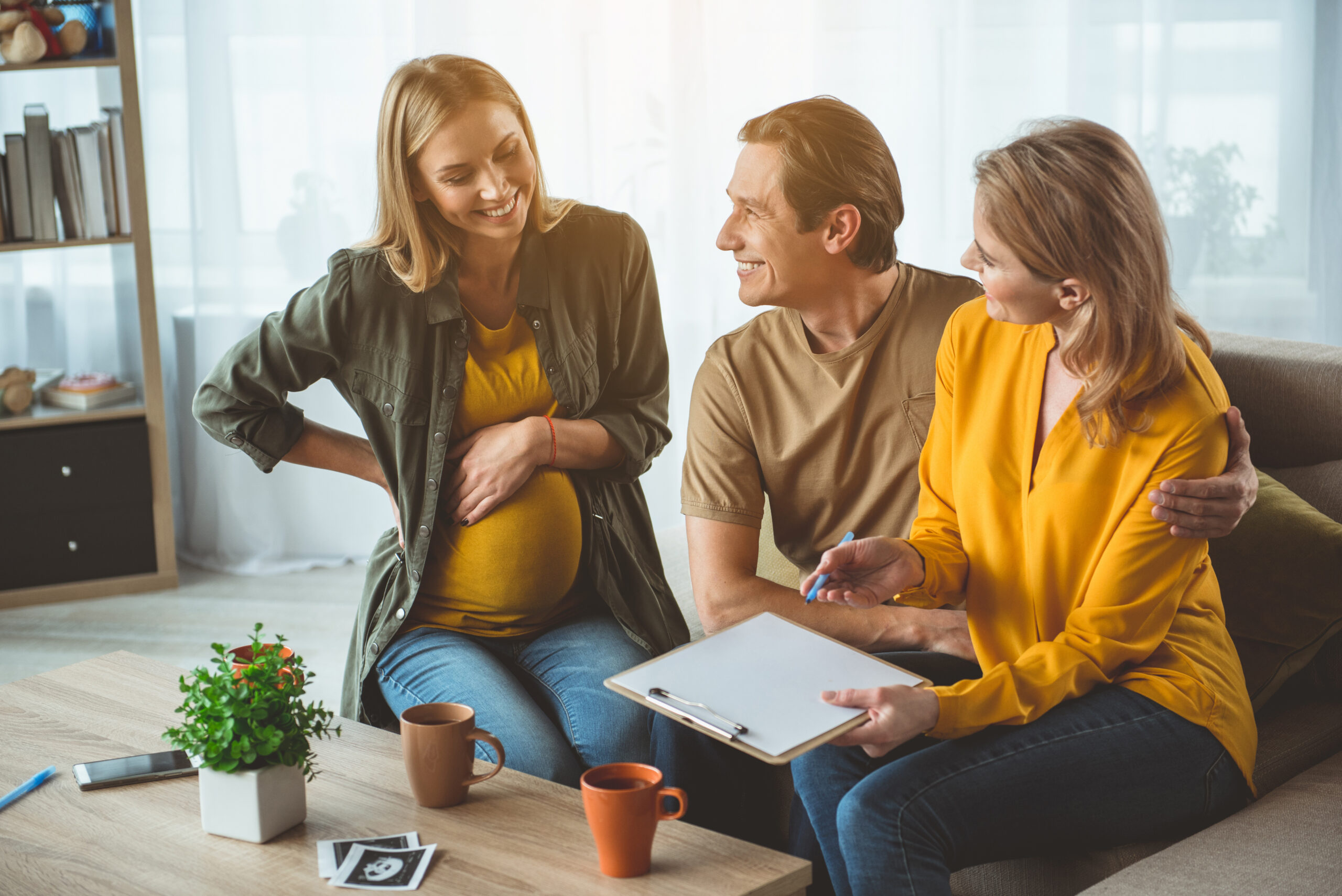 A pregnant woman sits with intended parents (male and female) look happy as they go over their surrogacy contract in a living room.