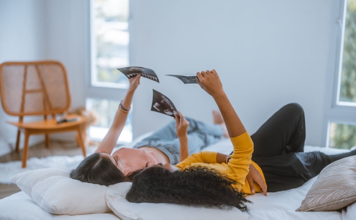 Two women lying on a bed at home, smiling and holding up ultrasound images while looking at them together in a bright, relaxed setting.
