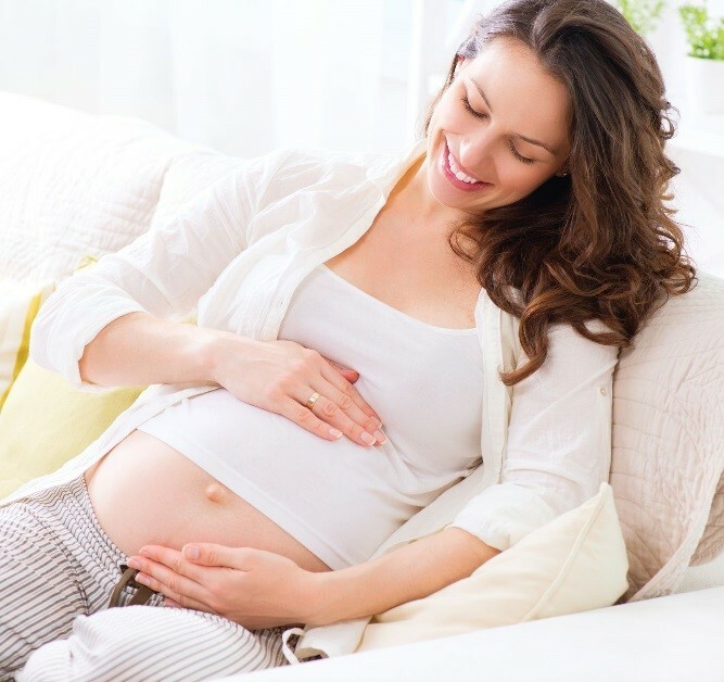 A smiling pregnant woman leans back on a couch looking down at her belly while cradling it with her hands.