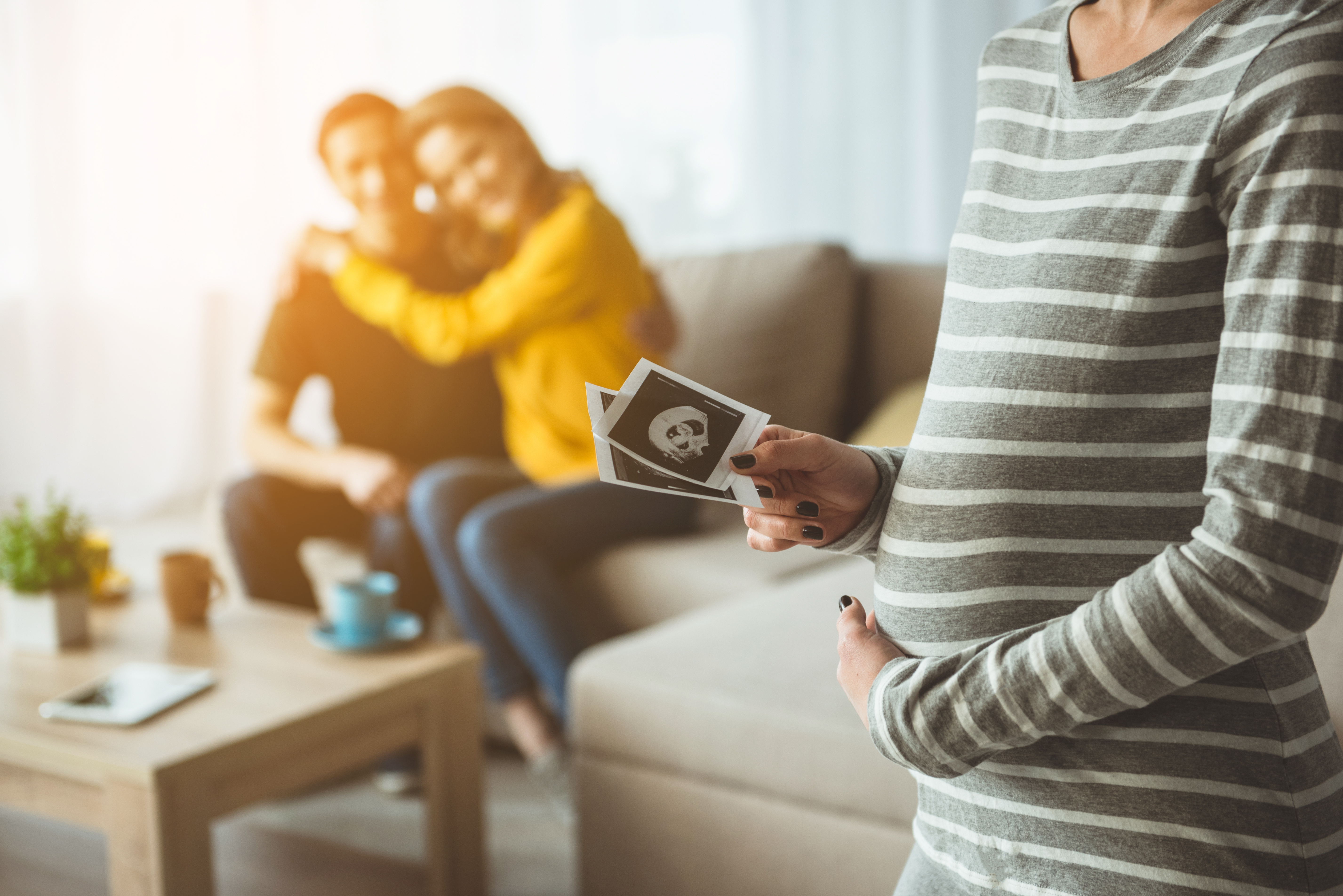 A surrogate mother cradles her pregnant belly, holding her ultrasound picture with the intended parents in hugging in the background on a couch
