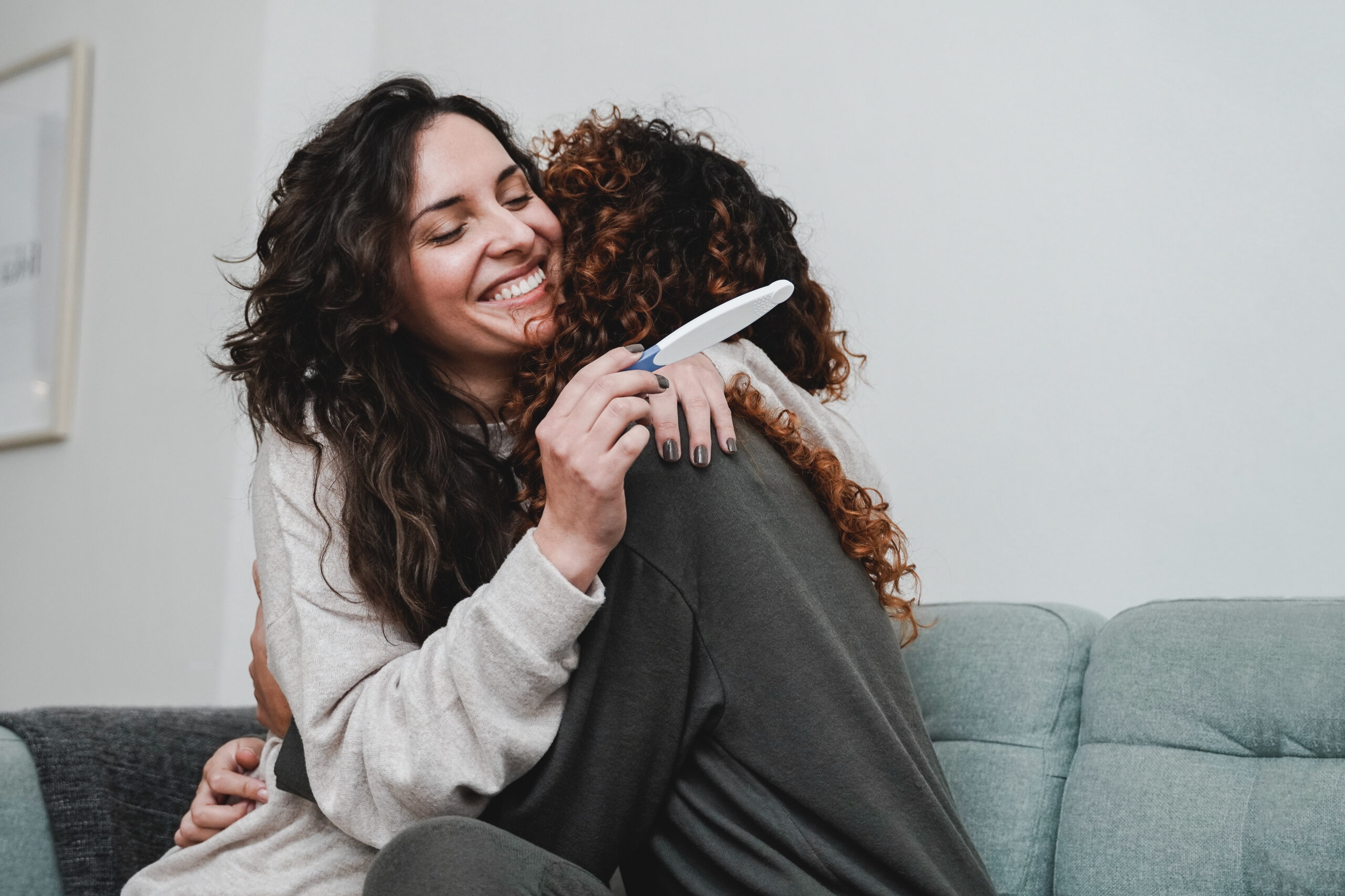 A surrogate mother embraces her friend, the intended parent, while joyfully holding a positive pregnancy test