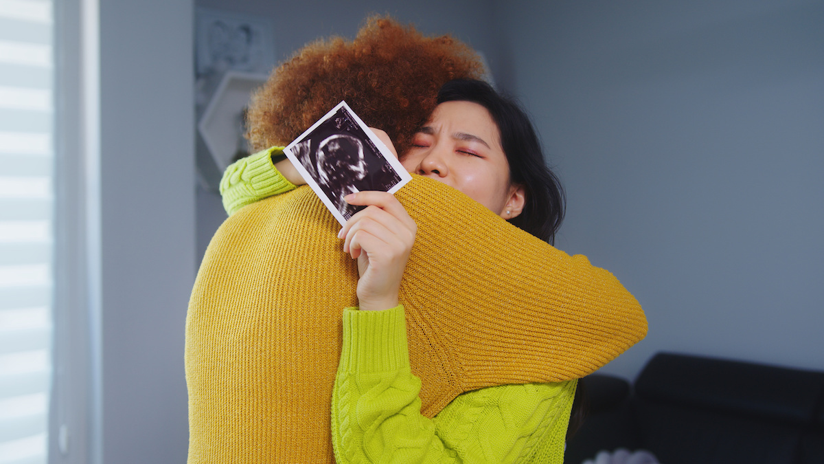 A woman is hugging a child while holding an ultrasound image. Both are emotional, as indicated by the woman's closed eyes and tearful expression. They are indoors, and the woman is wearing a bright yellow sweater.