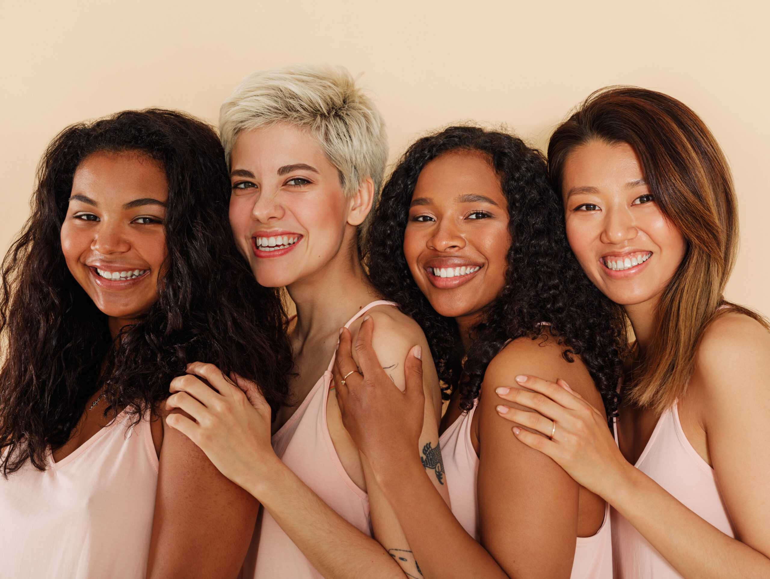 A group of four diverse, joyful women with beaming smiles, standing closely and embracing each other.