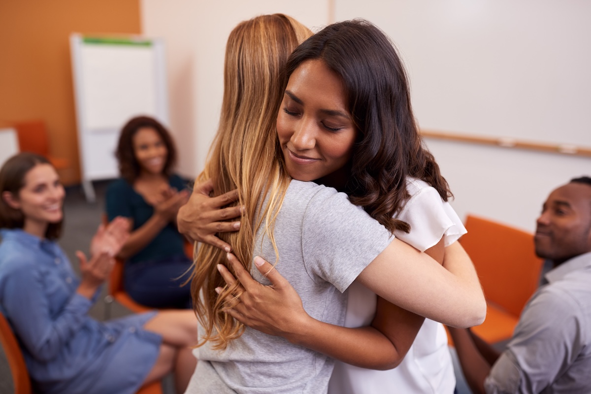 A tender moment within a support group setting, where one person is embracing another in a heartfelt hug while the rest of the group observes, some clapping in a gesture of support and community.