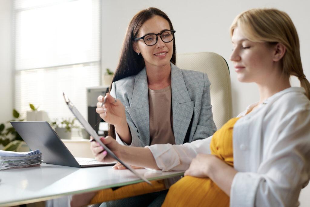Surrogacy agency coordinator reviews paperwork with a pregnant woman in a bright office, offering guidance as they look over a clipboard together.