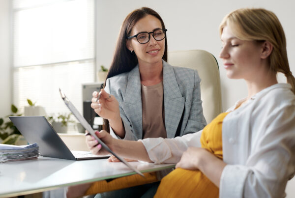 Surrogacy agency coordinator reviews paperwork with a pregnant woman in a bright office, offering guidance as they look over a clipboard together.