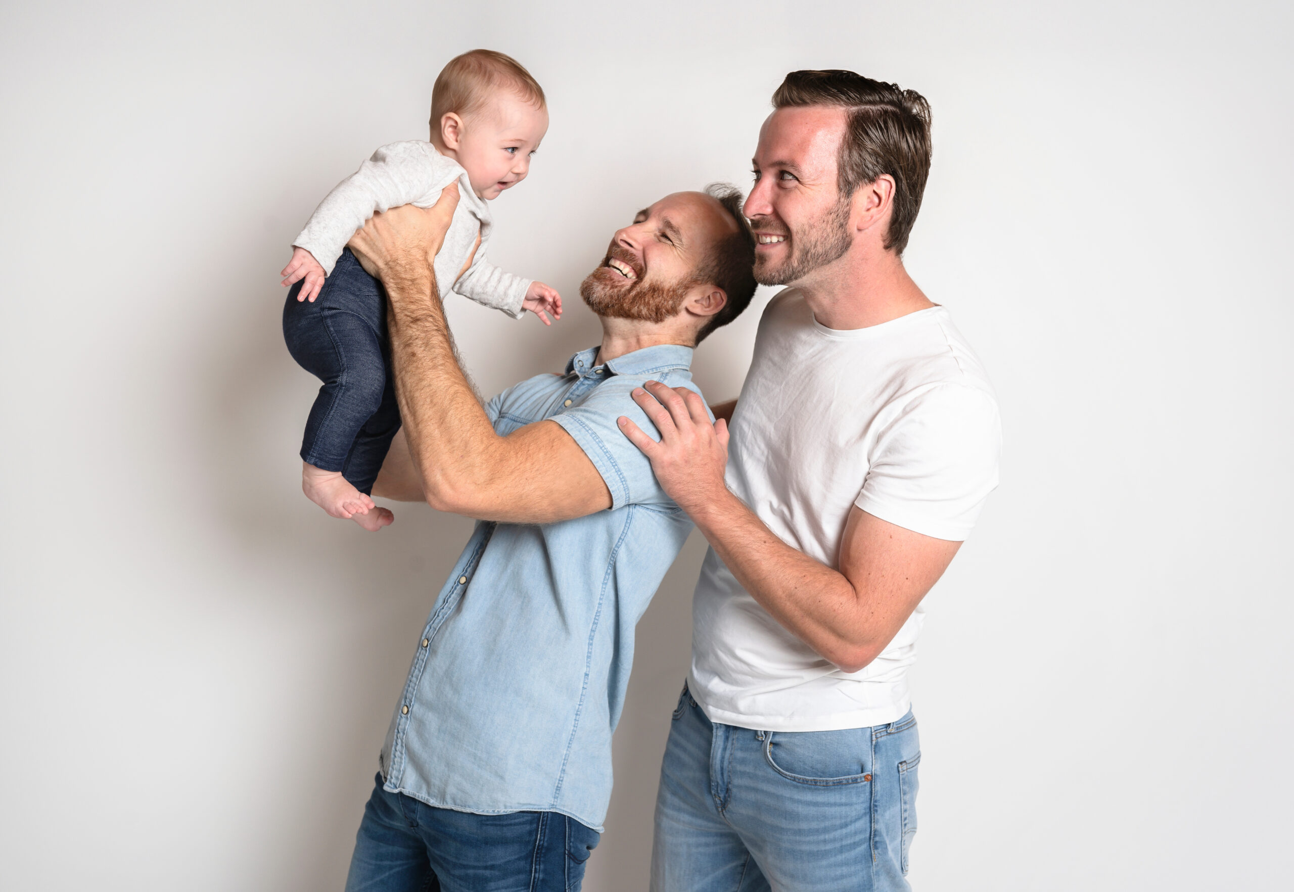 A gay couple looks up smiling at their child being held up by one of the men.