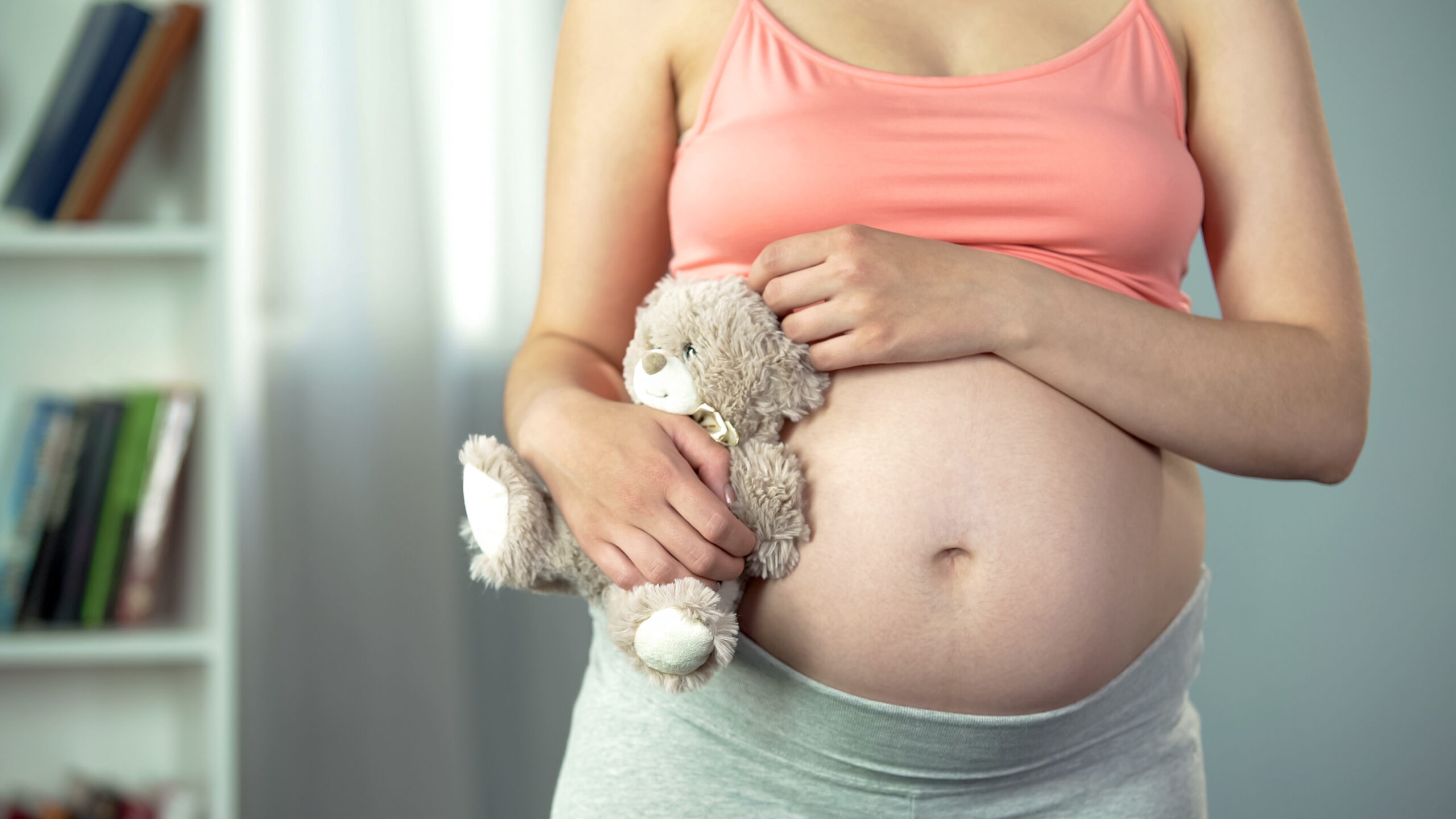 A pregnant woman wearing a crop shirt with her belly showing holds a teddy bear to her side.