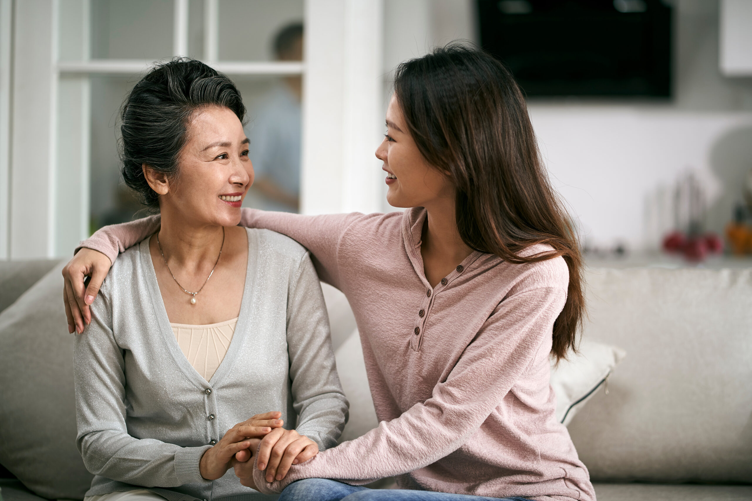 A mother and a daughter, sitting on a couch smiling at each other, with one woman placing her arm around the other in a gesture of affection.