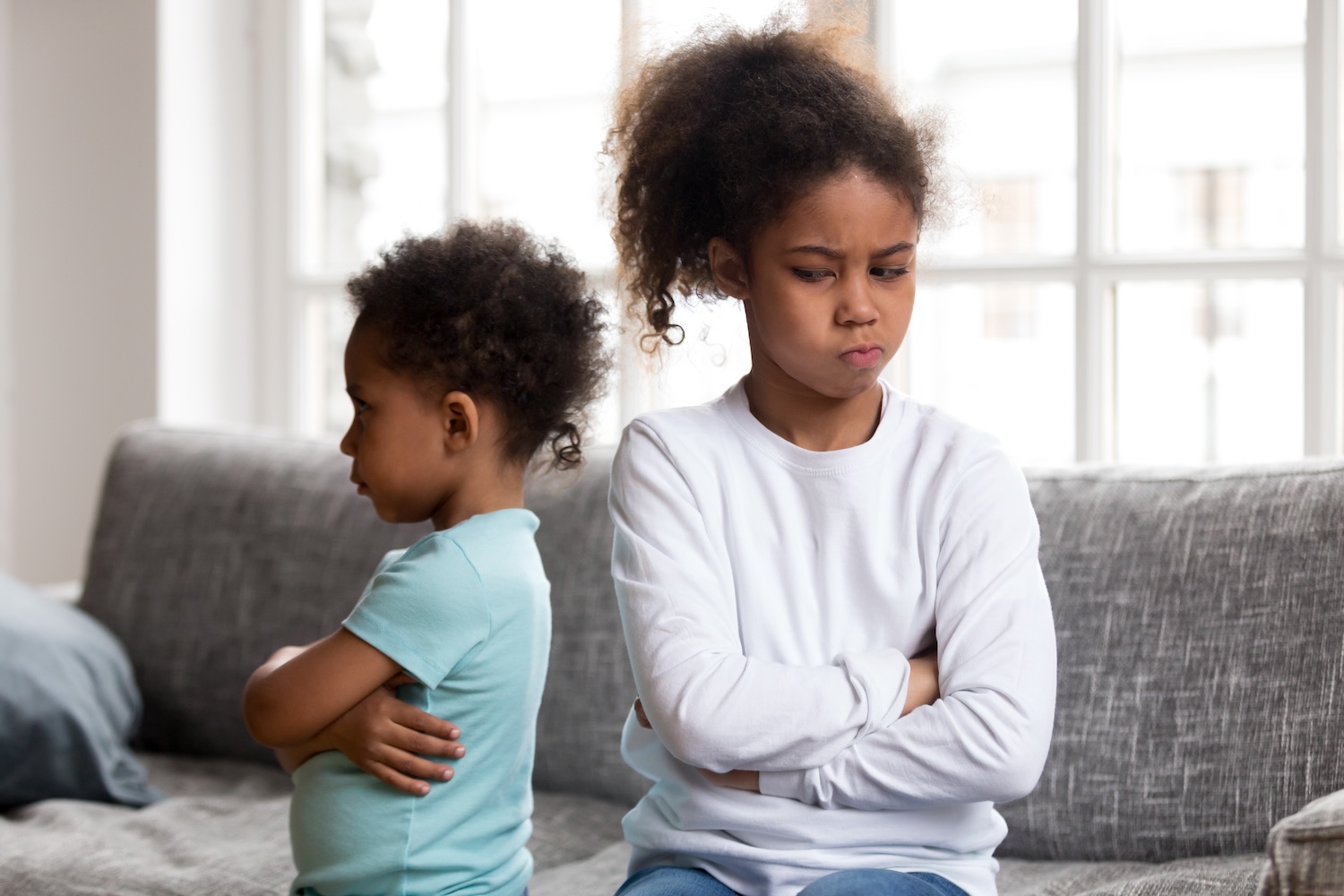 Two young children are sitting back-to-back on a couch with their arms crossed, both looking upset.