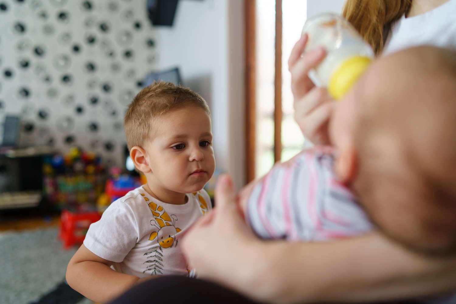 A young boy watches attentively as an adult feeds a baby with a bottle.