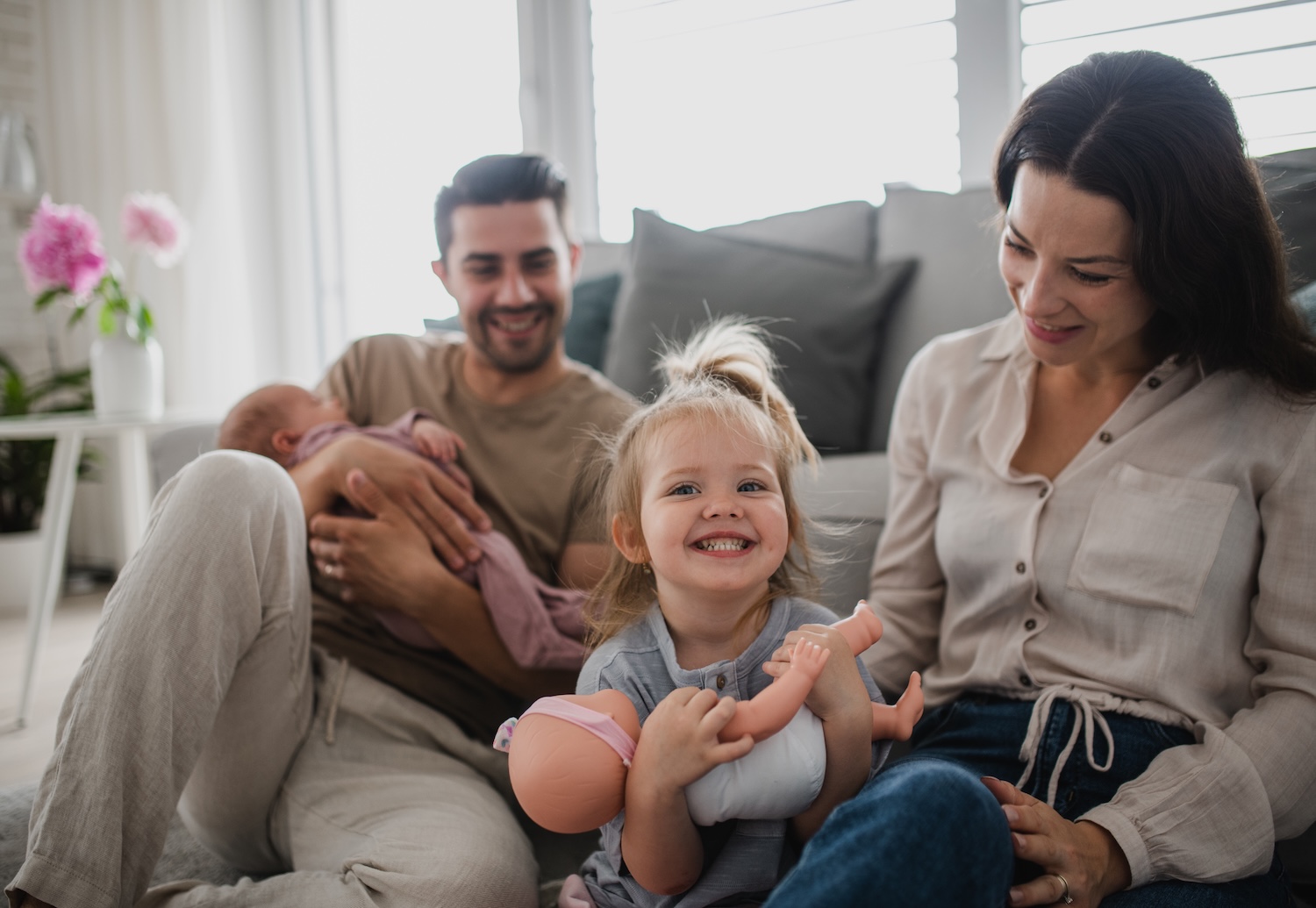 A smiling toddler holds a baby doll while sitting between her parents. The father cradles a newborn, and the mother lovingly looks at their daughter.