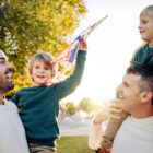 Two dads laughing with their young children outdoors at sunset, one child held on shoulders waving a kite in the air.