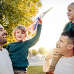 Two dads laughing with their young children outdoors at sunset, one child held on shoulders waving a kite in the air.