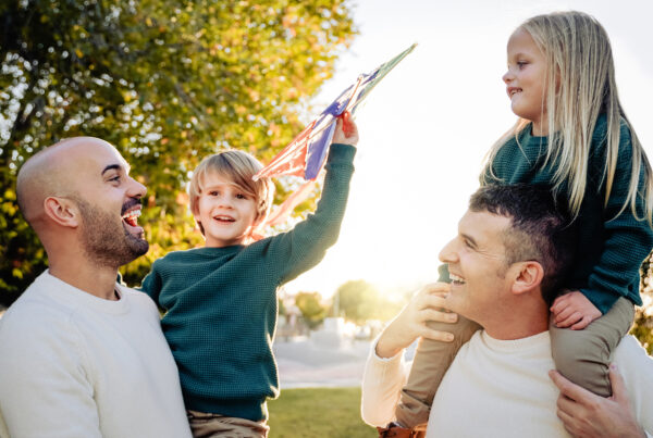 LGBTQ+ Surrogacy and Family-Building Options 7 Two dads laughing with their young children outdoors at sunset, one child held on shoulders waving a kite in the air.