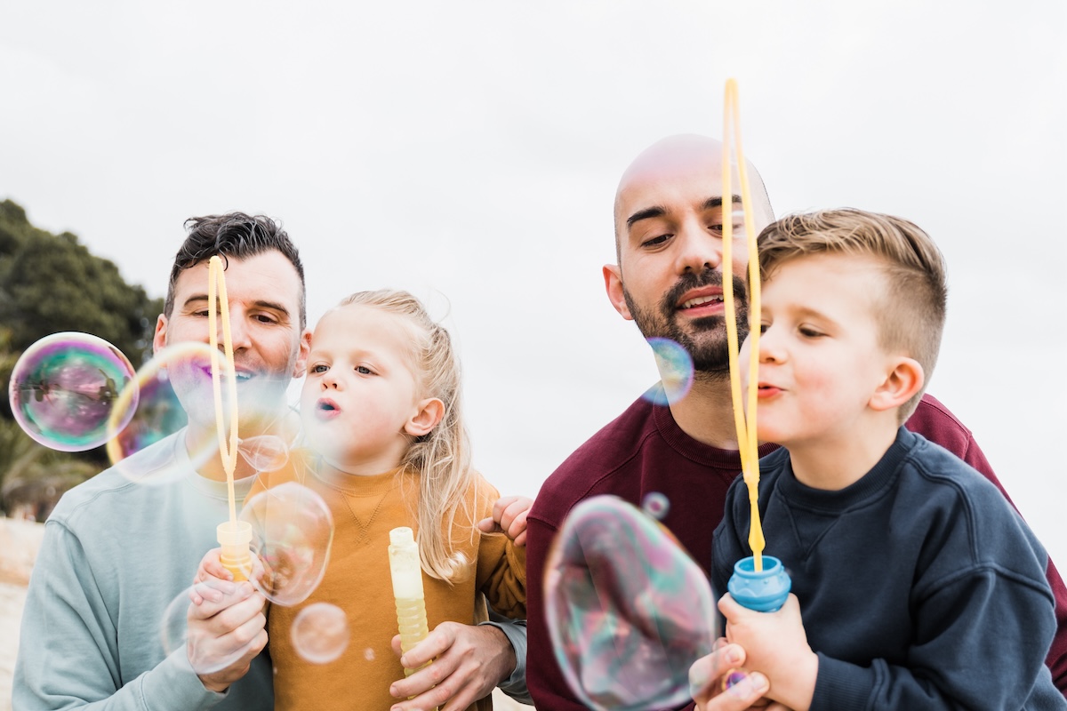 Gay parents and sons having fun with soap bubbles on the beach during summer vacation