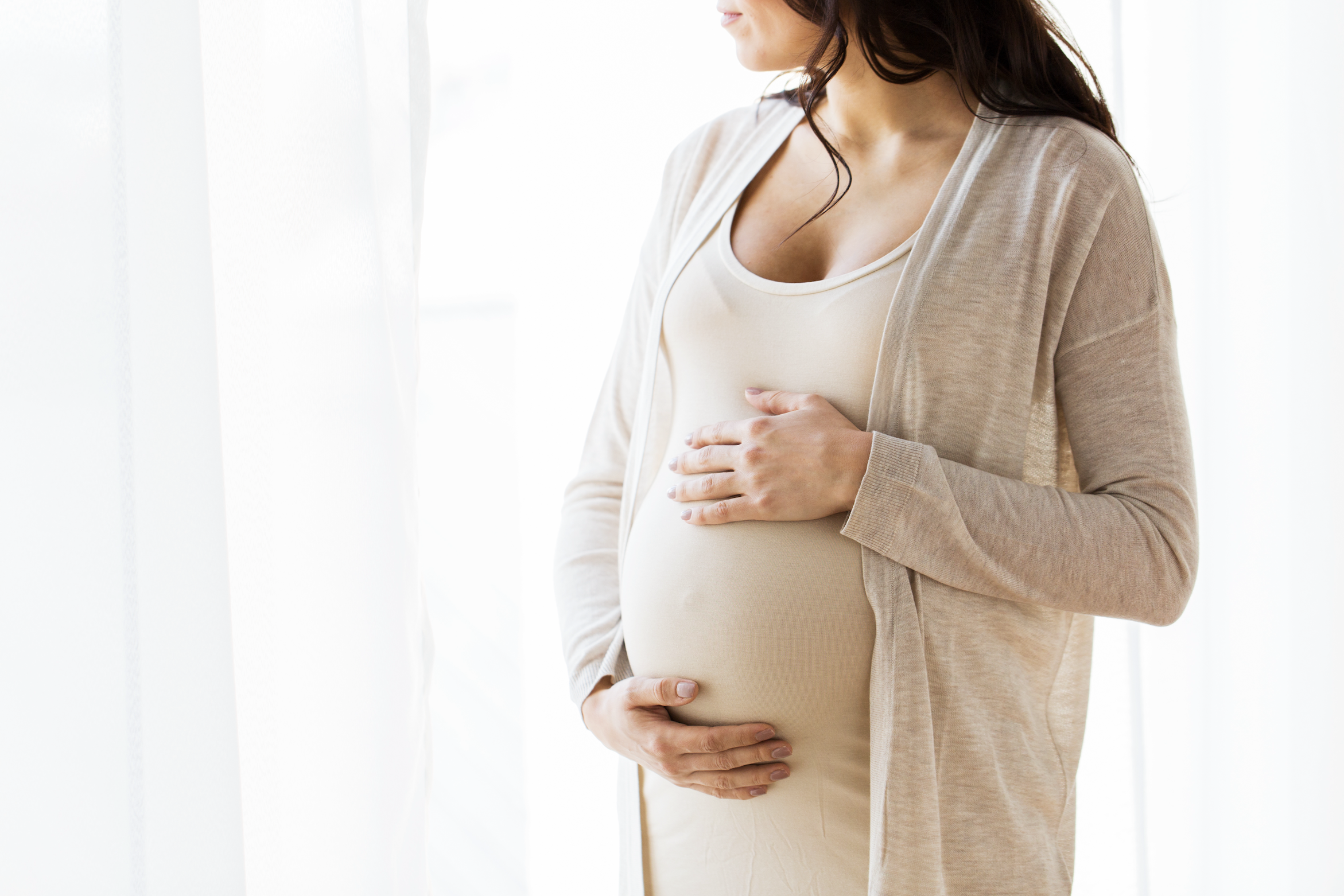 A close-up shot of a pregnant woman who is cradling her stomach while looking out the window to her right.