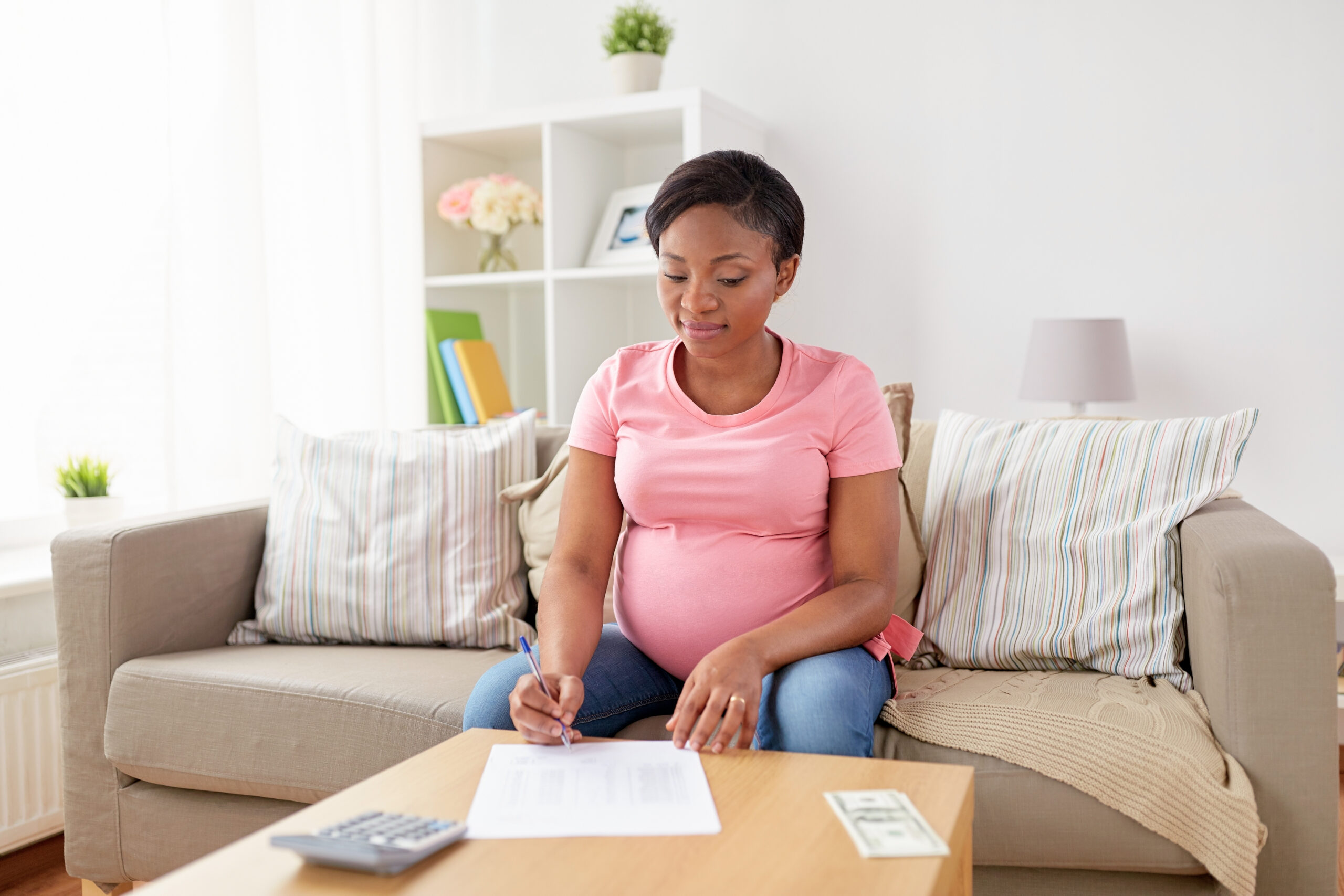 a pregnant woman sits on her couch working on her taxes with a calculator in front of her.