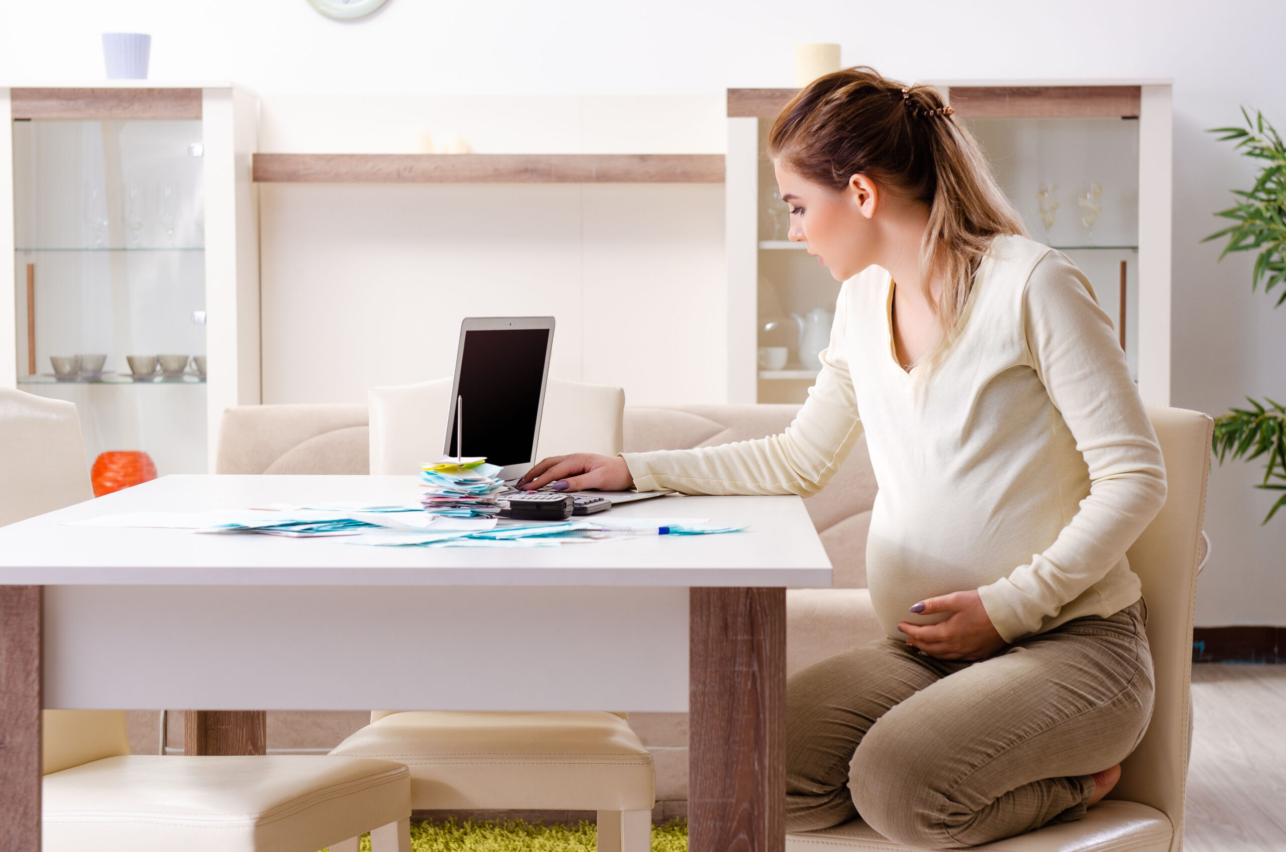 A pregnant woman sits at her desk with her laptop, looking through financial records and receipts.