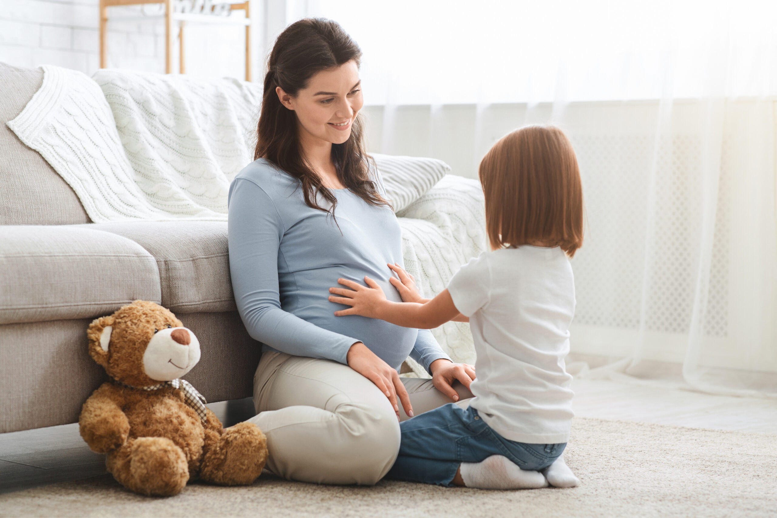 A surrogate mom in a cozy home setting, smiling as her daughter tenderly touches her belly.