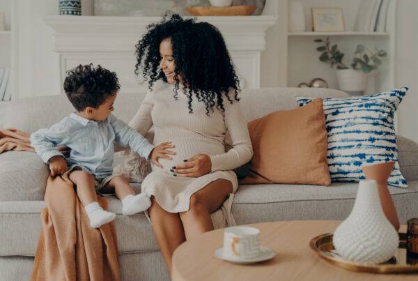 A stay-at-home surrogate mom sitting on a couch, engaging in a happy moment with her young son touching her belly.