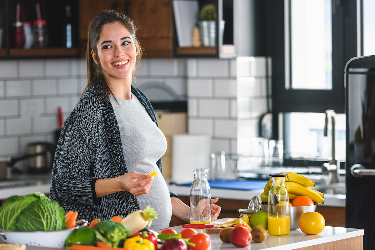 A pregnant woman prepares food in a modern kitchen, surrounded by fresh fruits and vegetables on the counter.