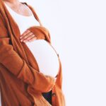 A surrogate mother standing and cradling her belly with both hands, wearing an burnt orange cardigan.