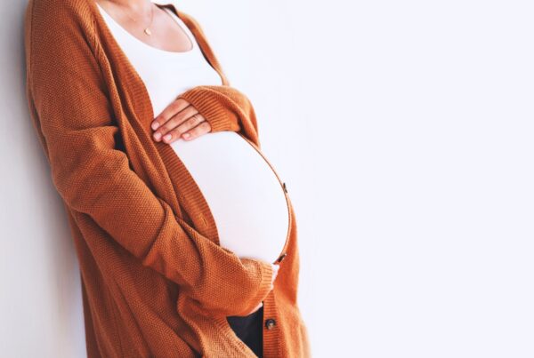 A surrogate mother standing and cradling her belly with both hands, wearing an burnt orange cardigan.