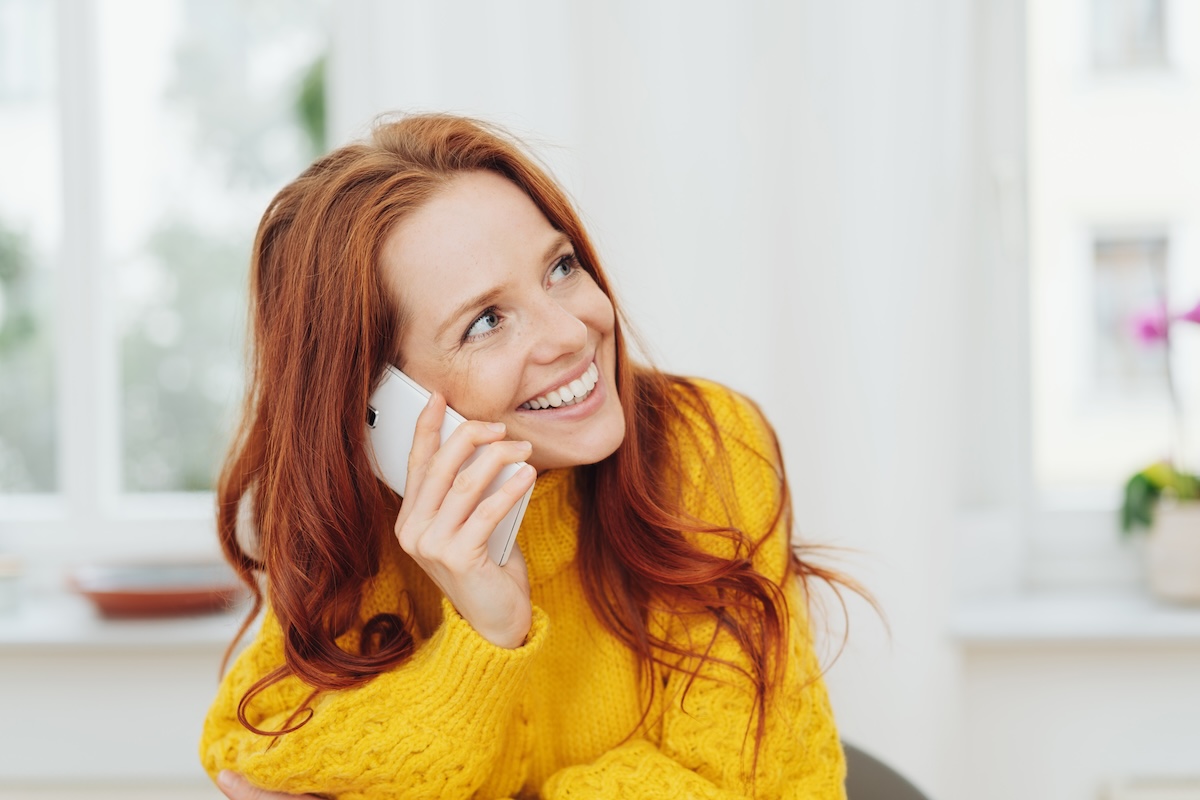 Woman with red hair talking on a smartphone, wearing a yellow sweater, and smiling while looking away.