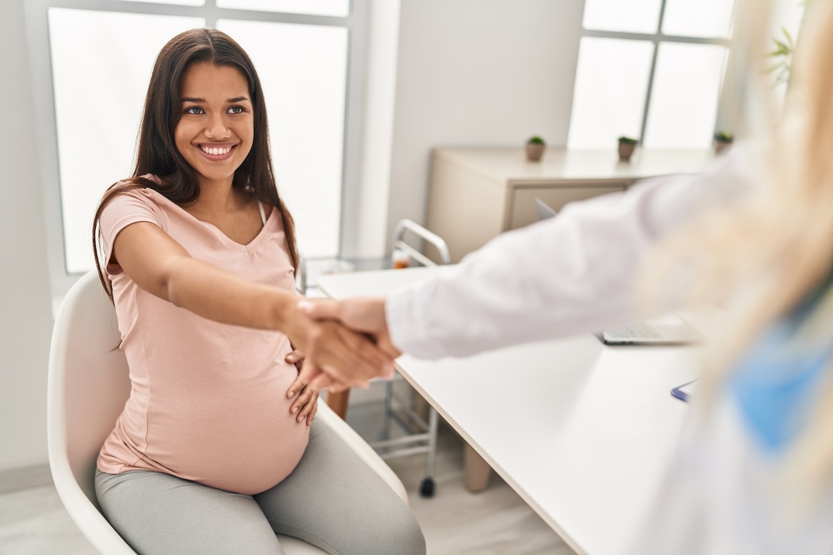 Pregnant woman shaking hands with another person across a table, smiling and touching her belly with her other hand.