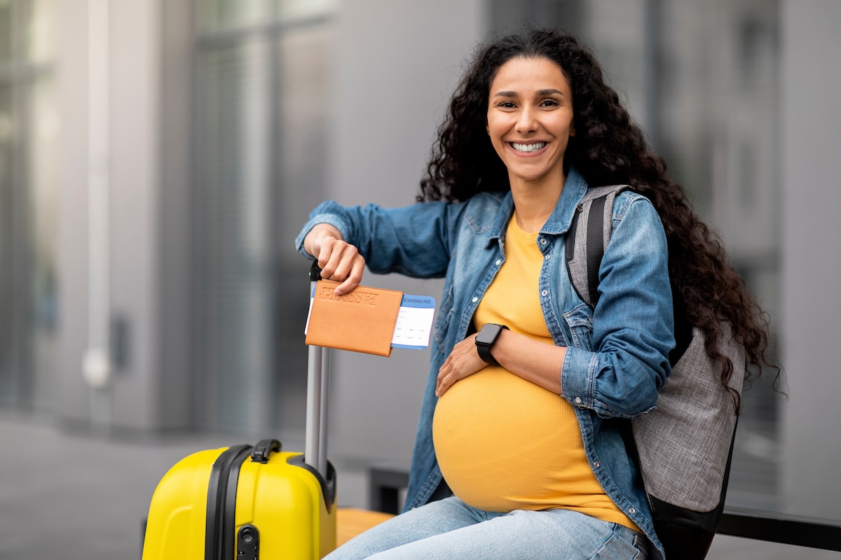 Smiling pregnant traveler with curly hair, wearing a denim jacket and sitting with a yellow suitcase, holding a passport and boarding pass.