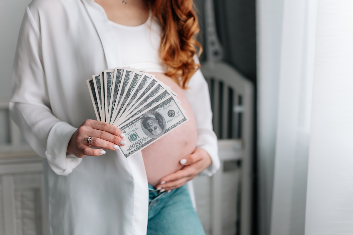 Pregnant person in white top holding a fan of dollar bills while touching their belly.