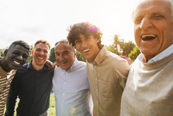 A diverse group of five men posing together, with expressions of joy and closeness. They are standing closely, some with their arms around each other, and appear to be laughing or smiling broadly.