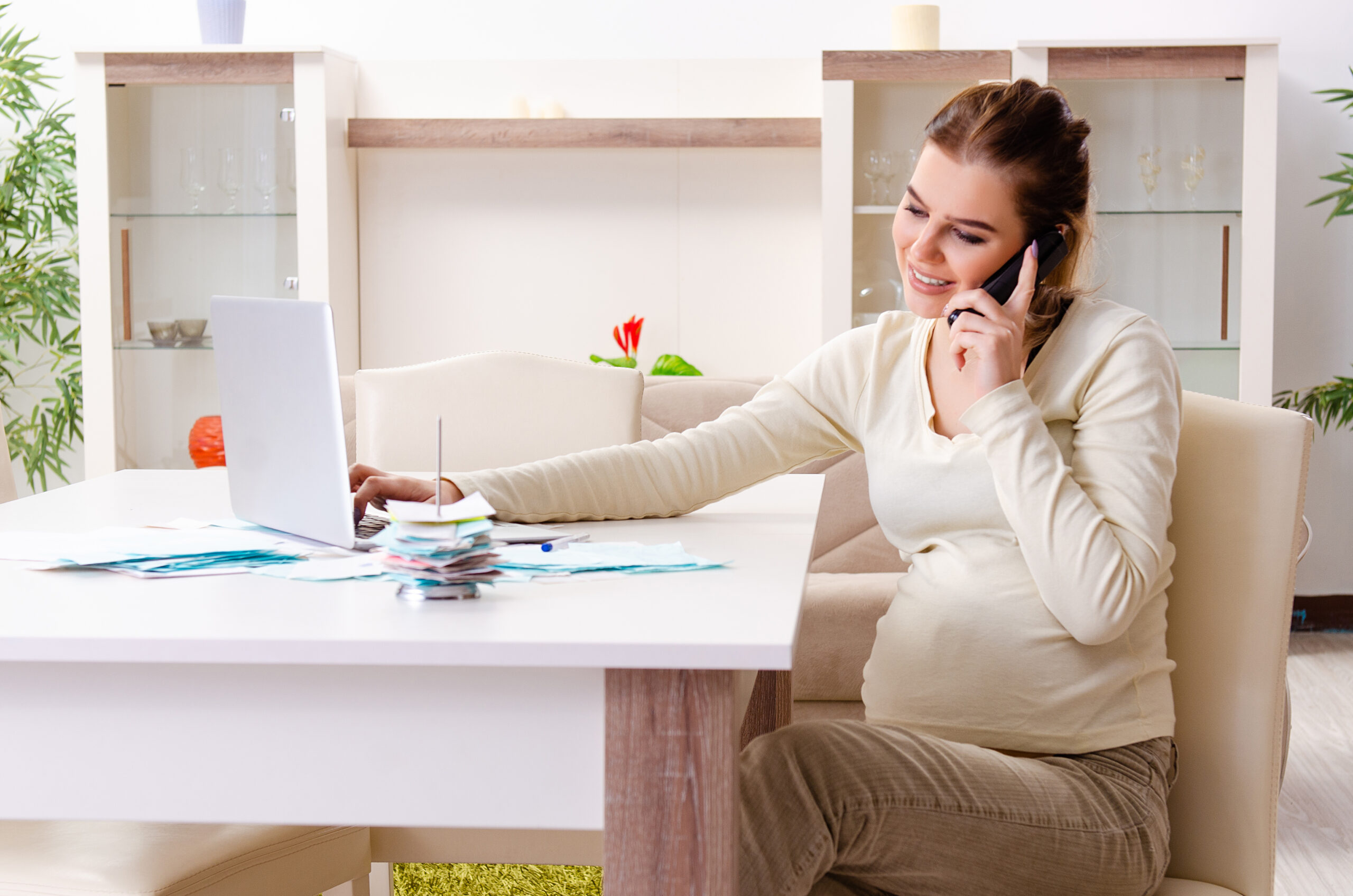A surrogate mother talking on the phone while using a laptop, with paperwork spread out on her table, discussing tax details with a financial advisor.