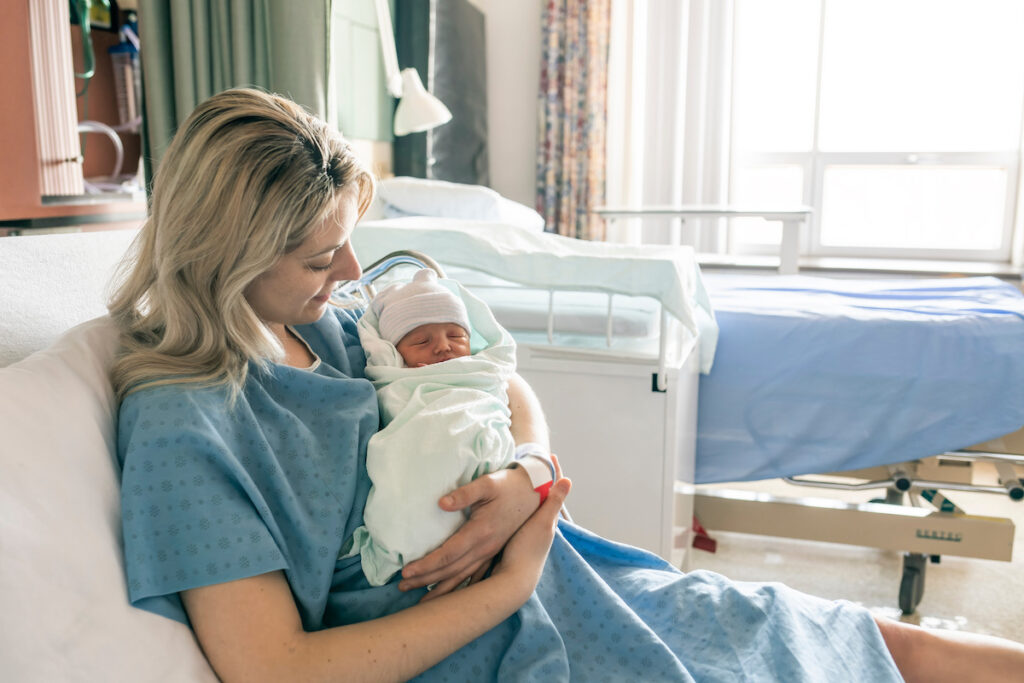 Una mujer en bata de hospital acunando a un bebé recién nacido en brazos, sentada en una cama de hospital con una ventana al fondo.
