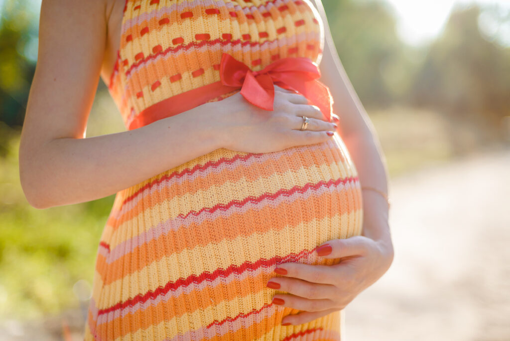 Pregnant woman in a bright, striped dress with a red ribbon above her belly, holding her bump with both hands, standing outdoors.