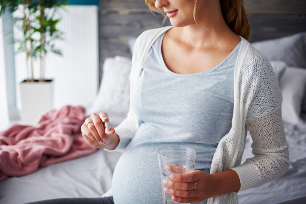 孕妇舒适地坐在床上，手里拿着一颗药丸和一杯水，准备服药。