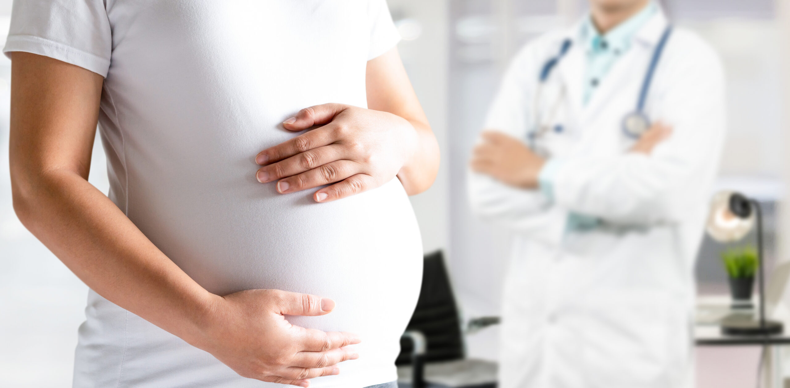 Pregnant woman standing while touching her belly with a doctor in the background.