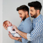 兩個身穿條紋襯衫、微笑的男子慈愛地抱著並看著一個新生嬰兒。