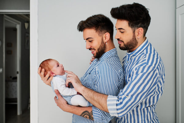 Two smiling men in striped shirts lovingly holding and looking at a newborn baby.