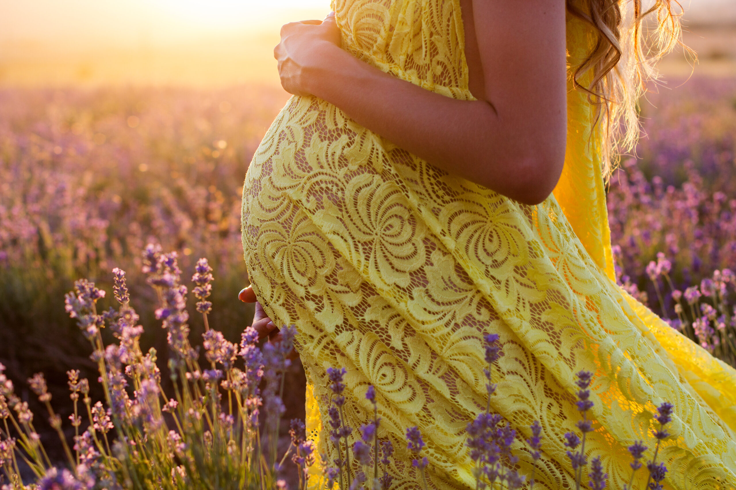 Pregnant woman in yellow sundress standing in a field of lavender flowers at dusk.