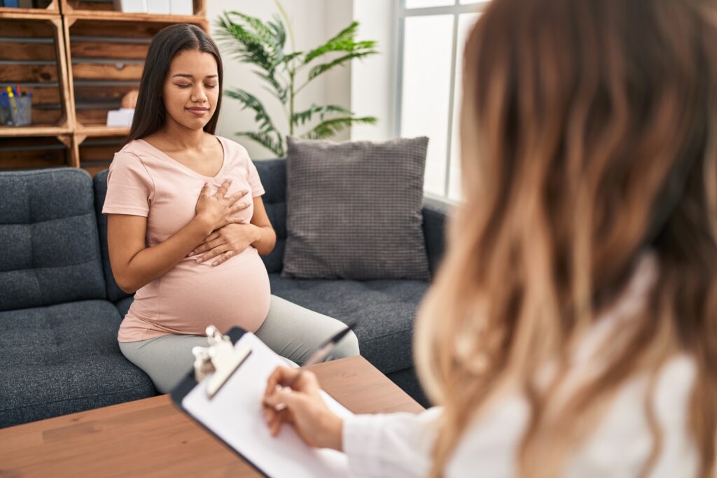 孕妇坐在沙发上，双手放在胸前，接受心理健康专家的咨询并做笔记。