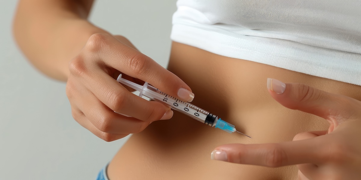 Person holding a syringe close to their abdomen, preparing to inject, wearing a white shirt.