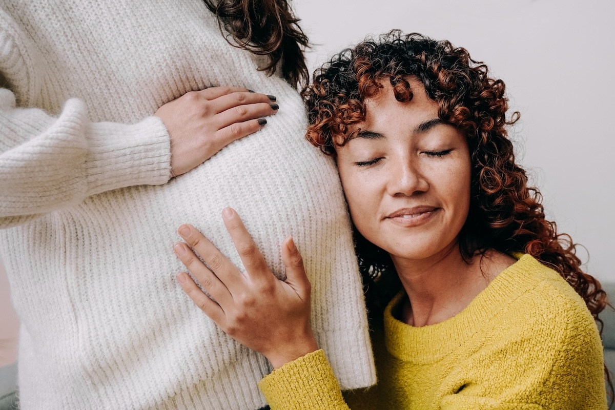 A smiling person with curly hair leaning their head against a pregnant woman's belly, hands gently touching the belly, both wearing sweaters.